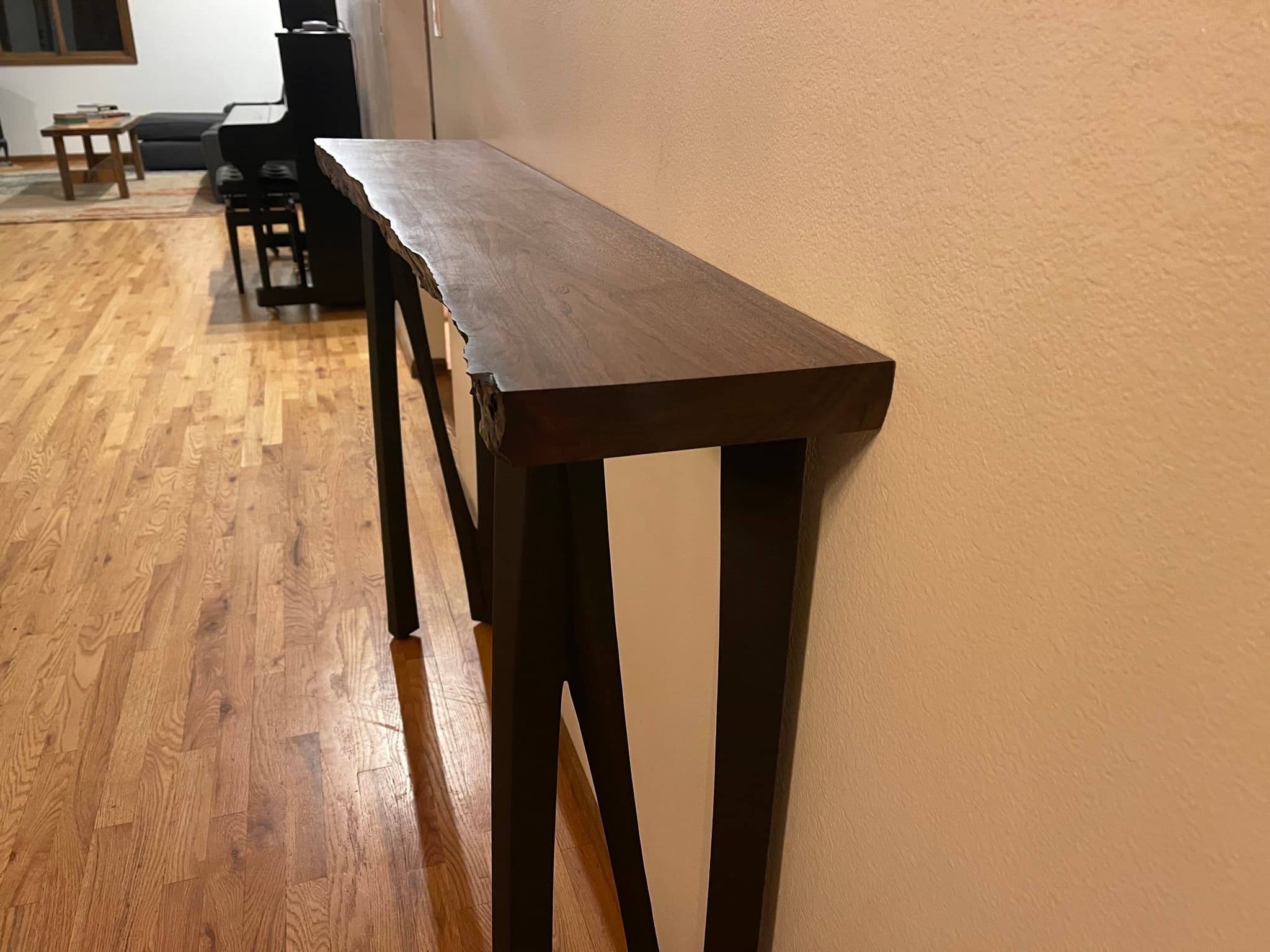 Walnut live-edge console table installed along a hallway wall, low three-quarter angle showing the slab profile and four splayed blackened steel A-frame legs over a hardwood floor.
