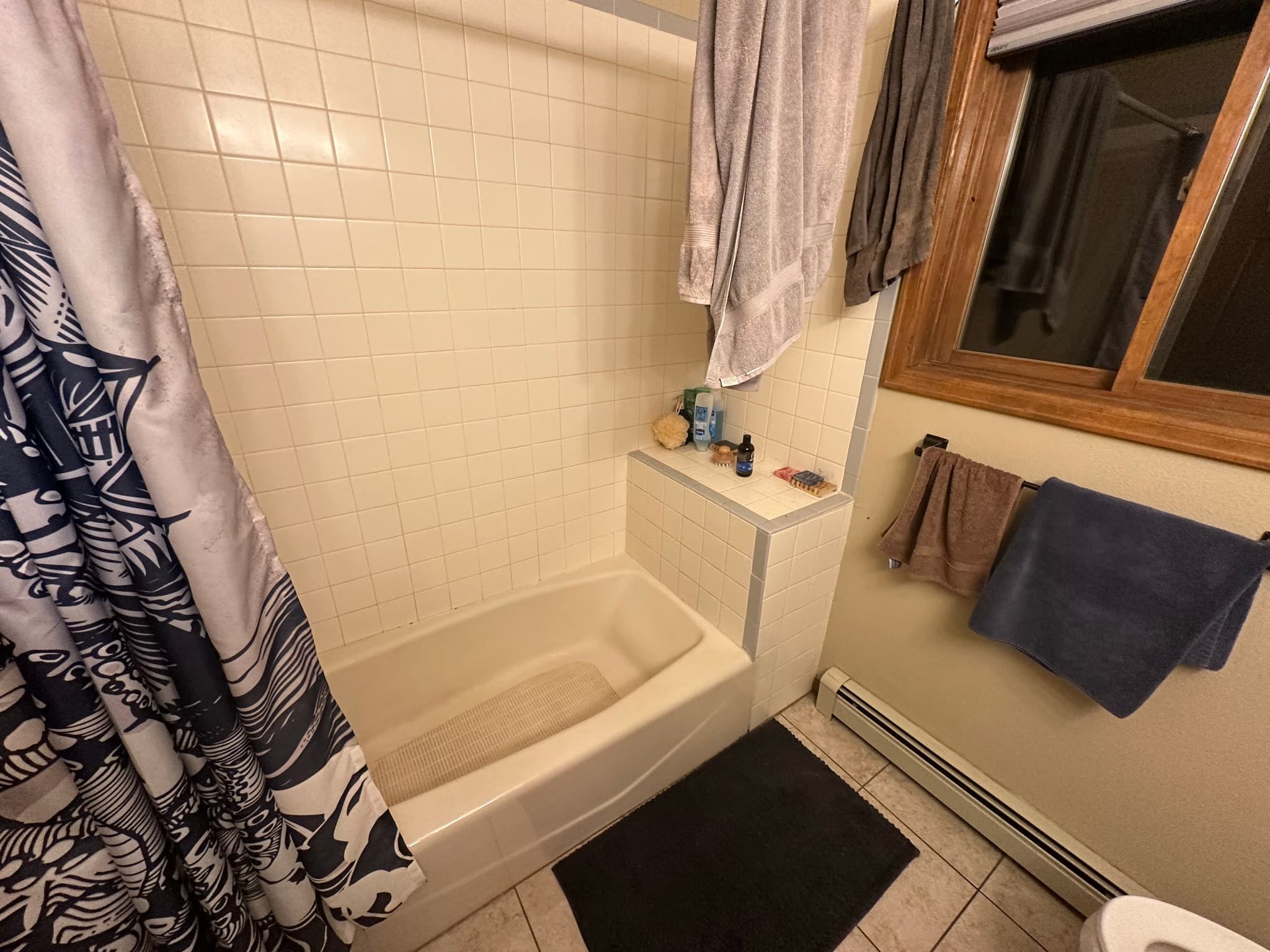 The original bathroom before work began: small cream wall tile, beige built-in tub, patterned shower curtain, oak-framed mirror, baseboard heater along the wall.