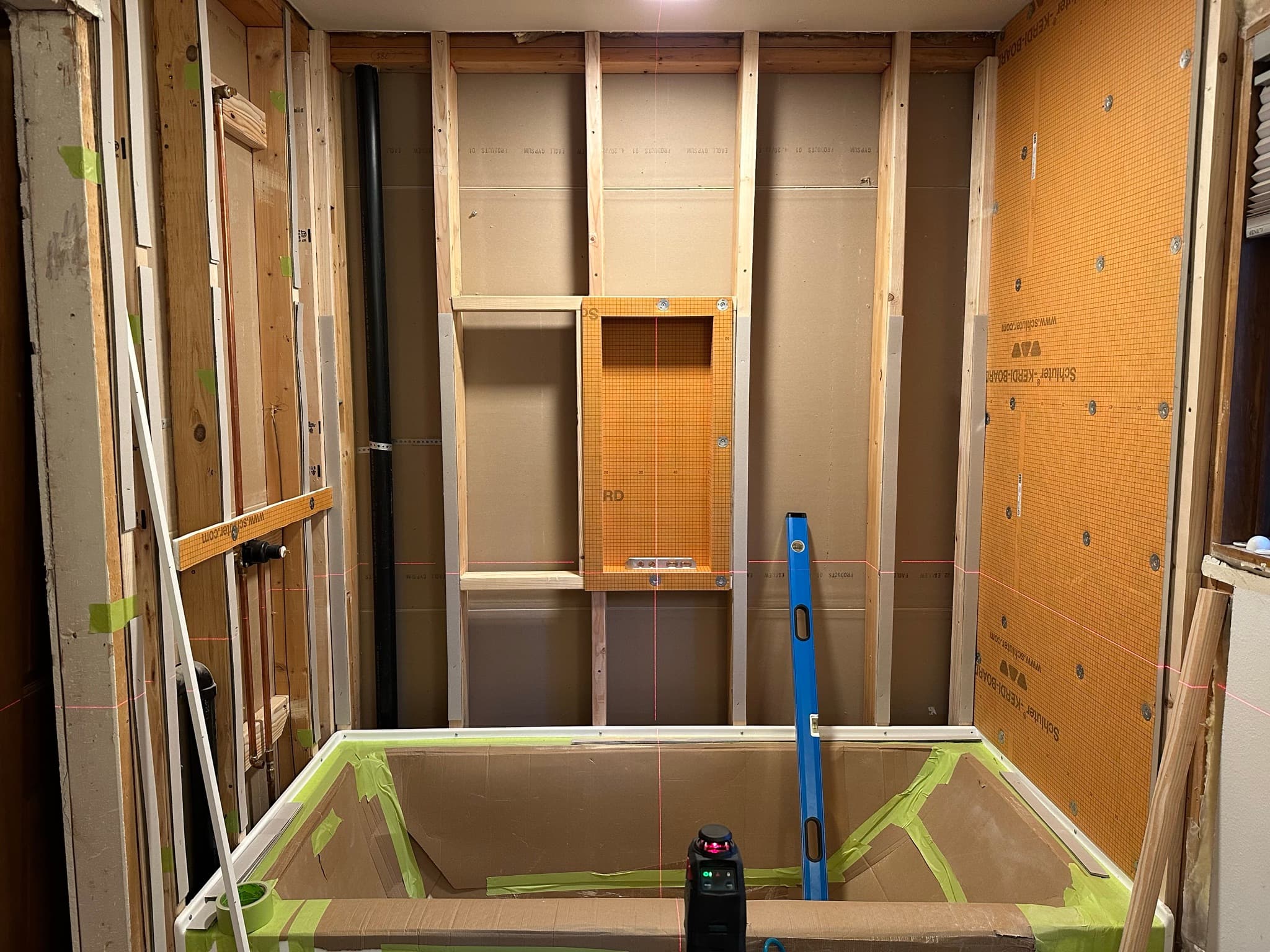 Tub apron protected with painter's tape and cardboard, new wood blocking added in the studs for the niche and grab bars, orange Schluter Kerdi-Board starting to go up on the right wall.
