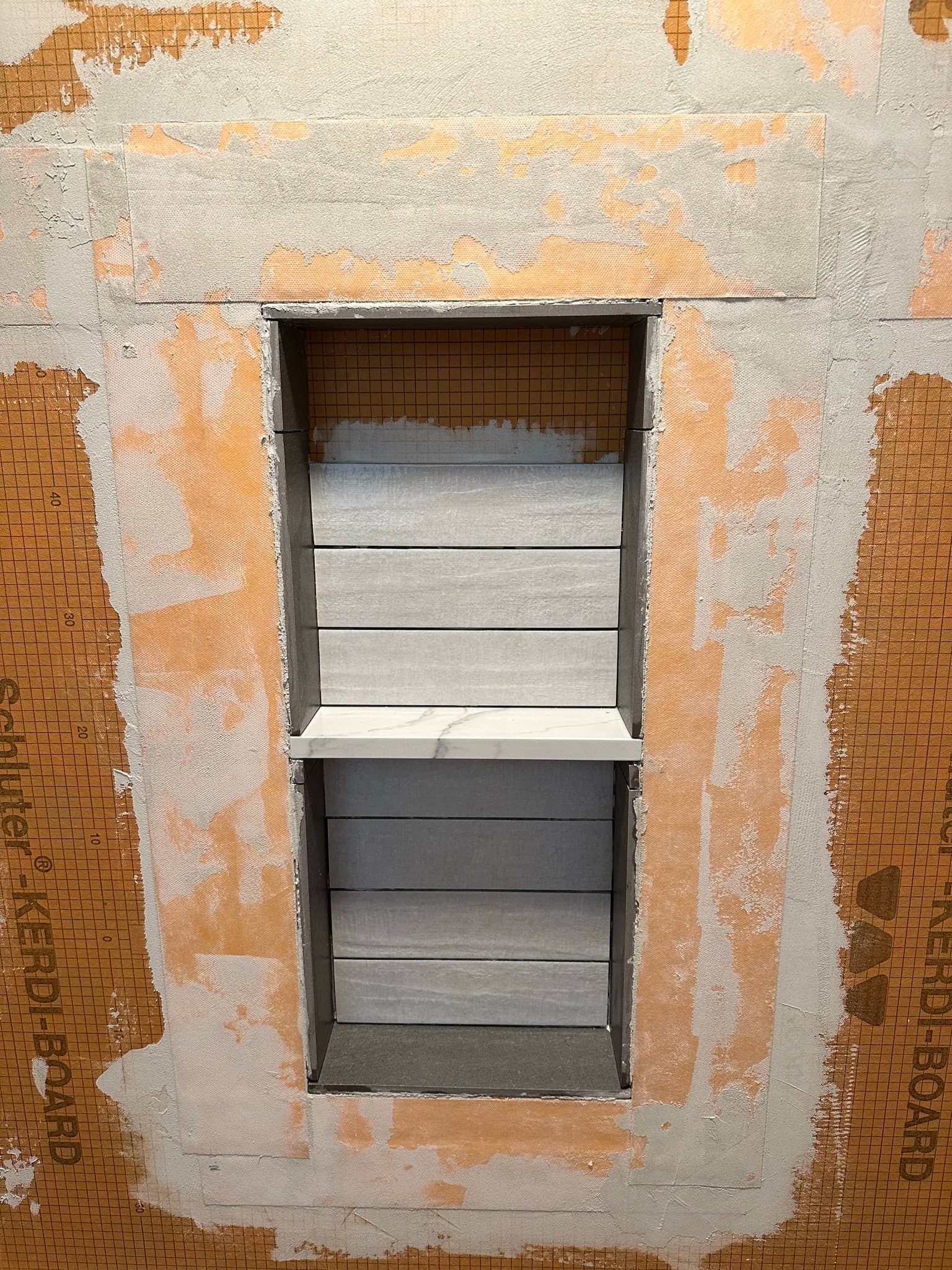 Two-compartment niche tiled out: stacked light-gray tile on the back wall, marble shelf splitting top and bottom, surrounded by un-tiled Kerdi-Board.