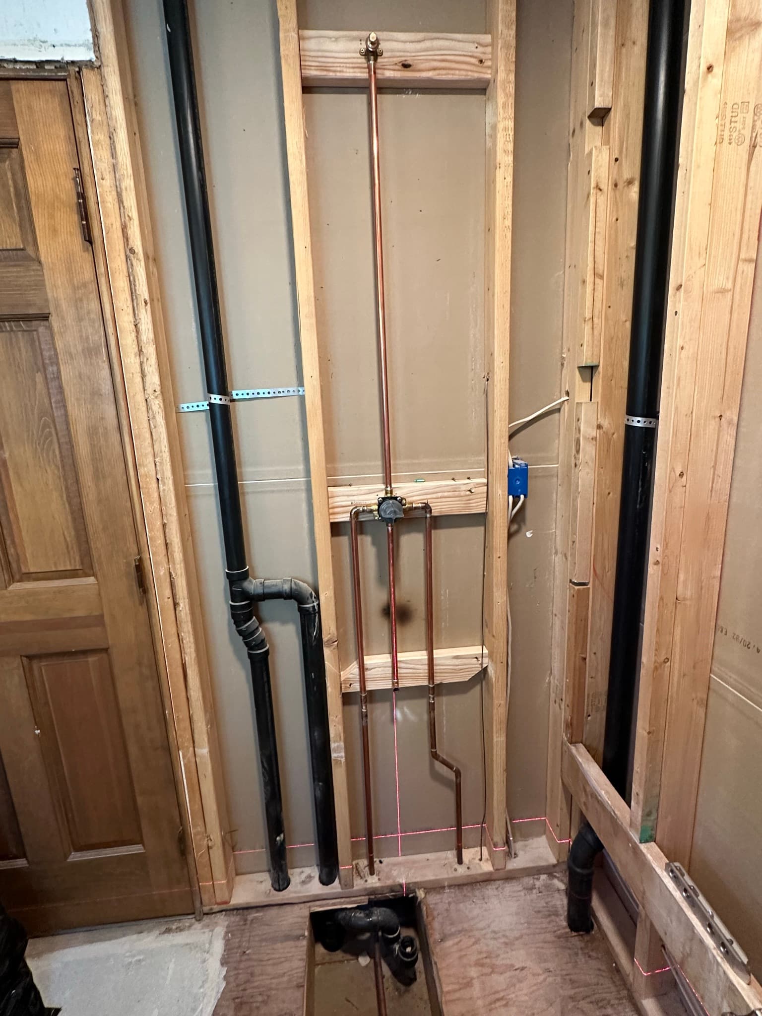 Re-plumbed shower wall: new copper supply lines running up to a brass mixing valve mounted between studs, ABS DWV roughed to the right, a red laser level line across the studs.
