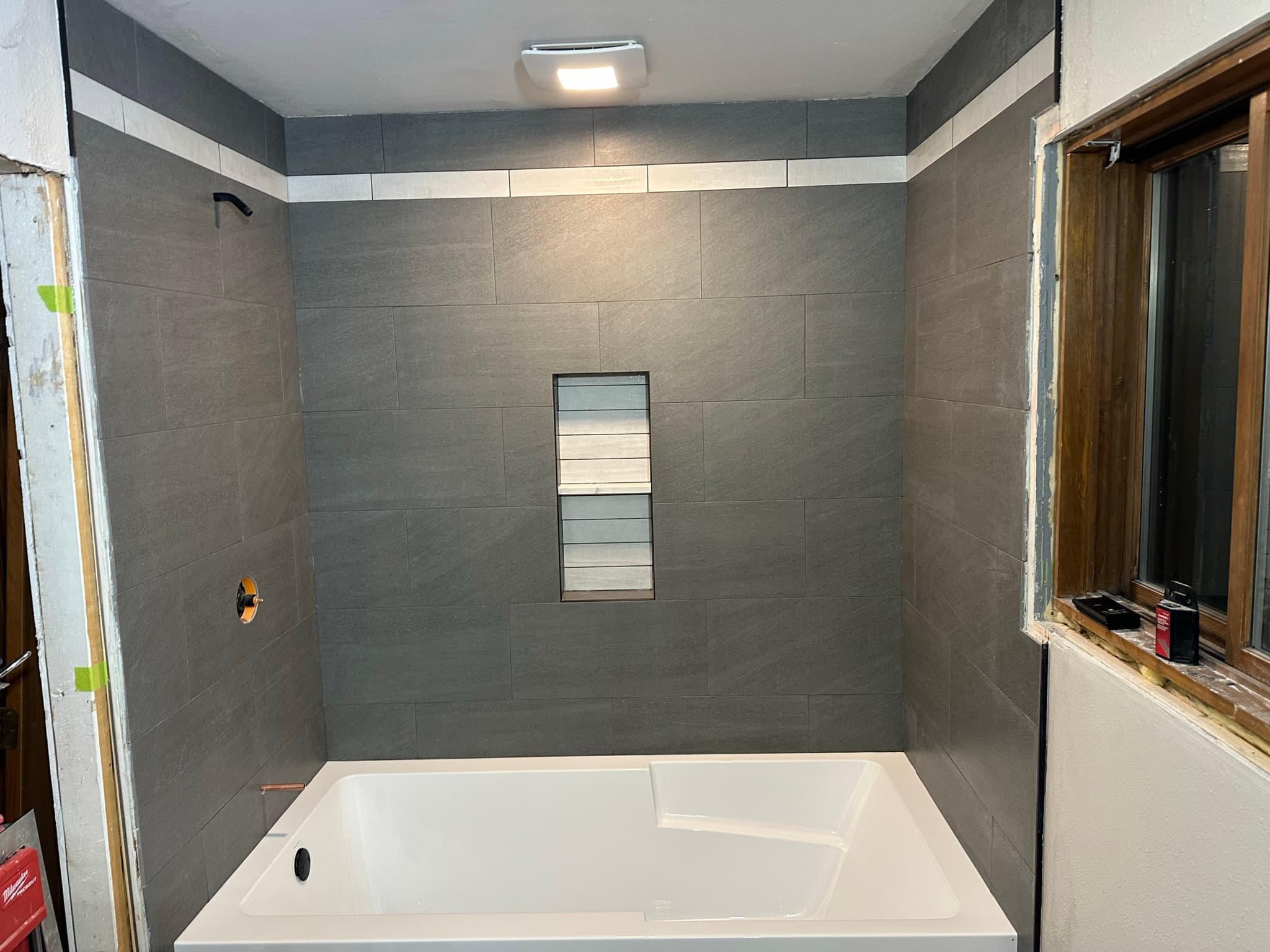 Finished tub surround: three walls of dark gray porcelain with a thin marble accent stripe near the ceiling, niche visible center-back, black shower valve trim, integrated apron tub below.