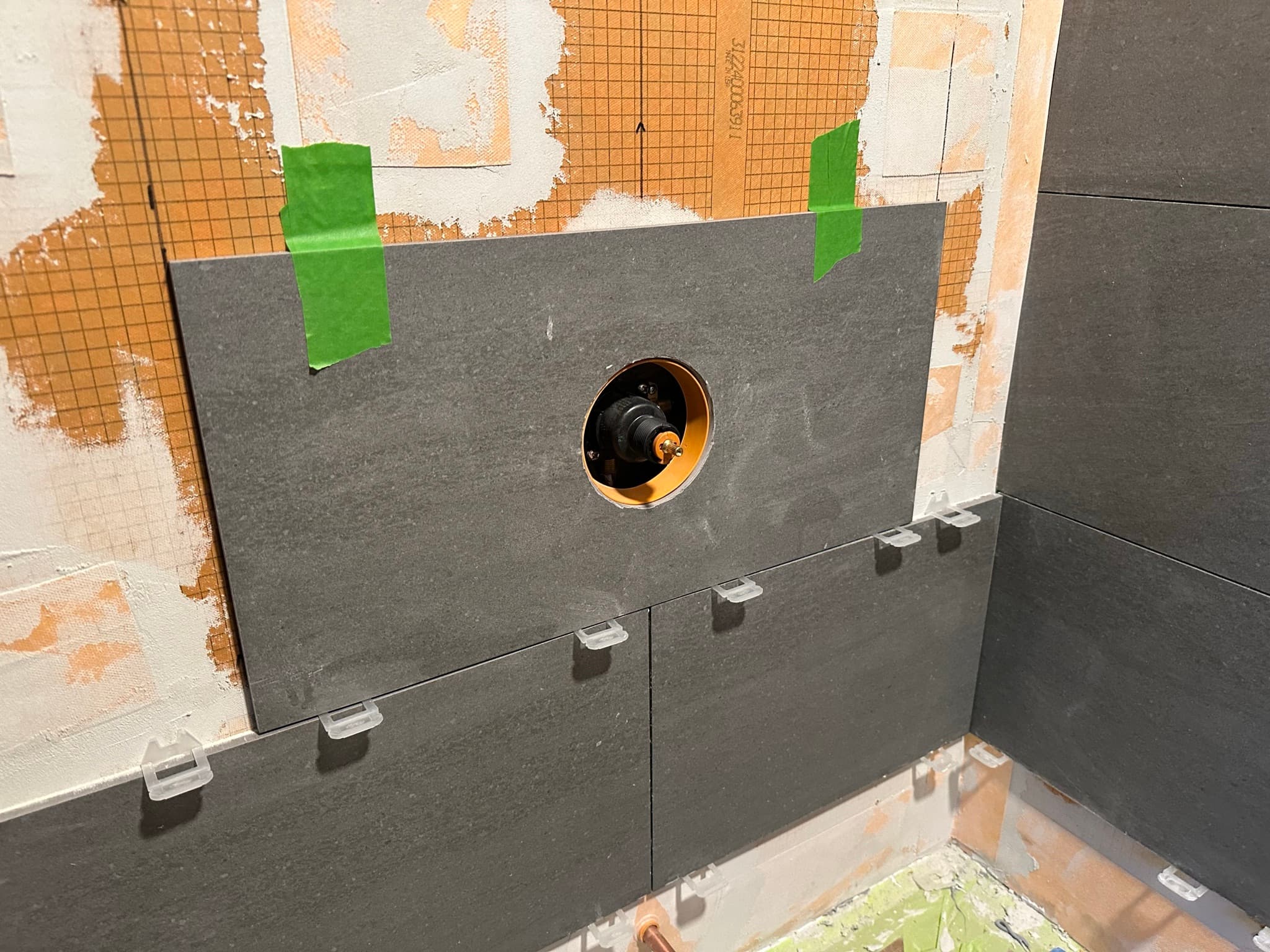 Close-up of a dark gray tile with a clean circular cutout around the brass mixing valve, leveling clips visible along the bottom edge.