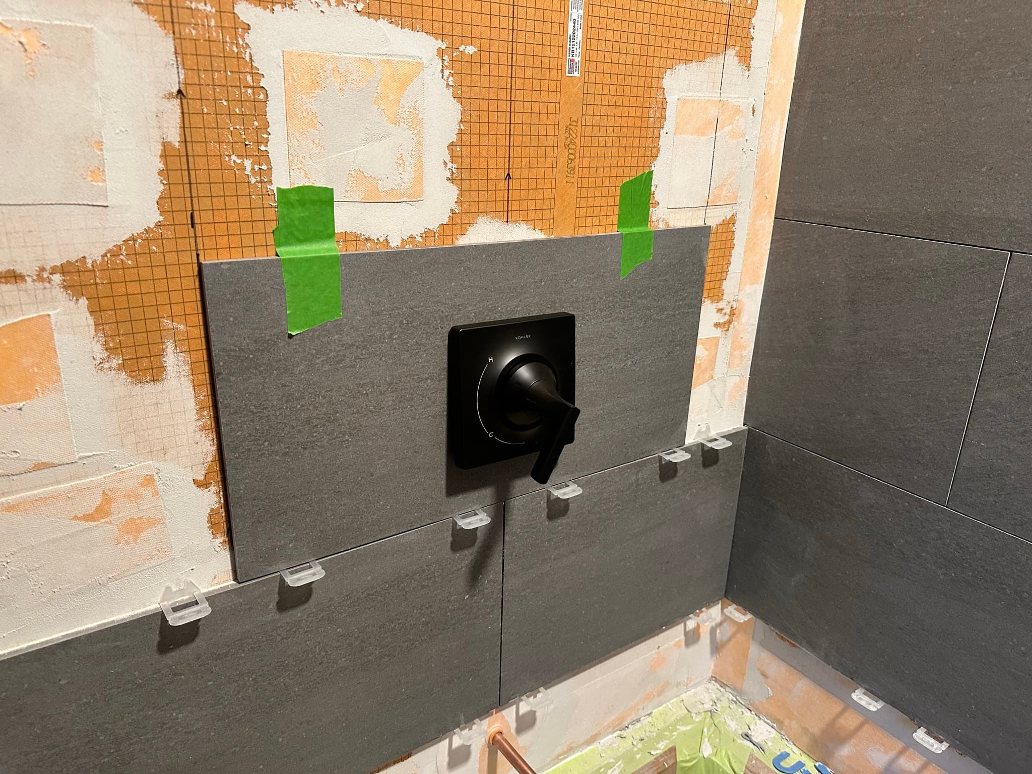 Matte black shower valve trim seated against the same dark gray tile, leveling clips still visible at the joints below.