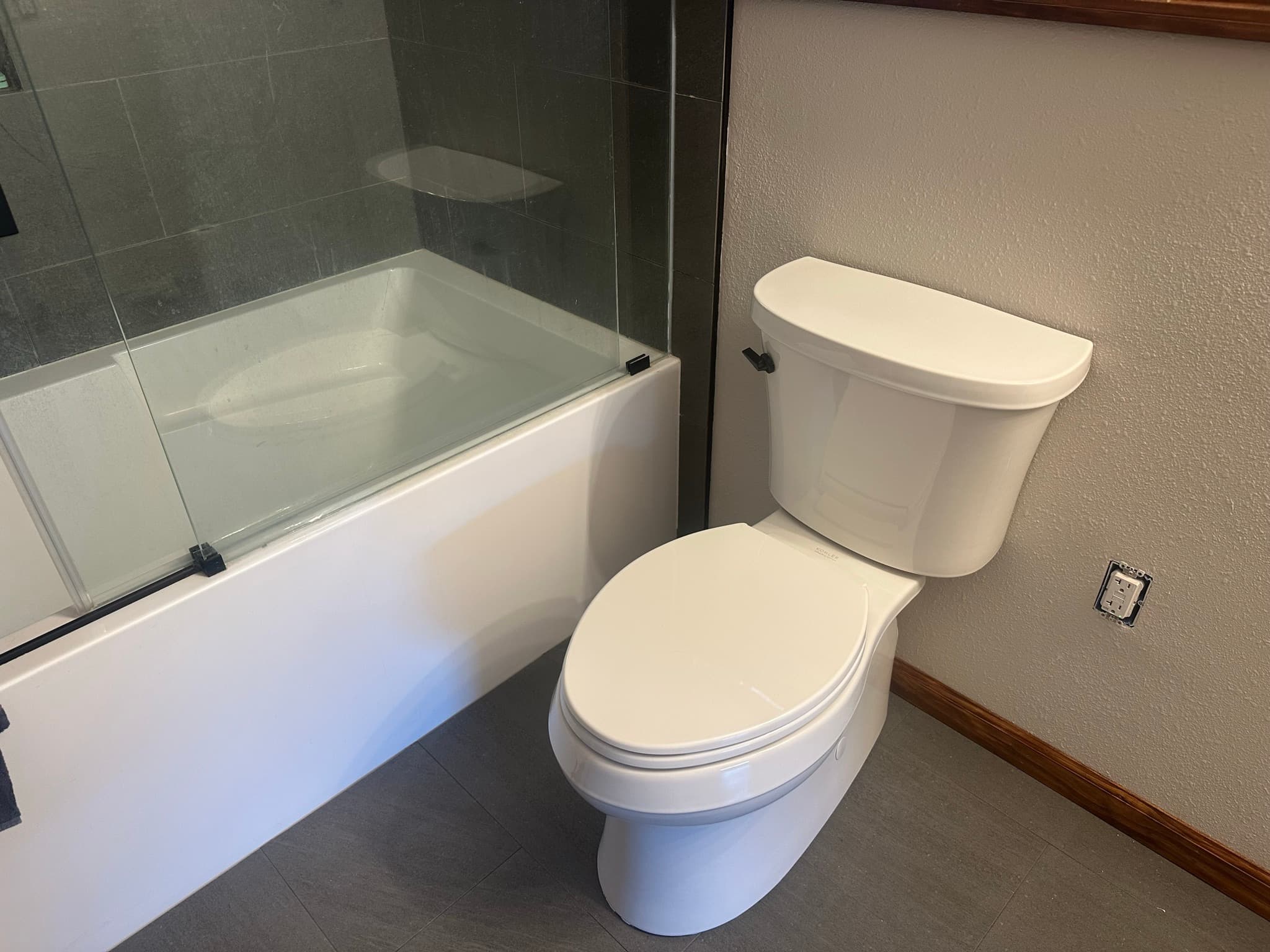 New low-profile toilet installed next to the glass-doored tub on the dark gray floor tile, baseboard heater grille visible against the wall.