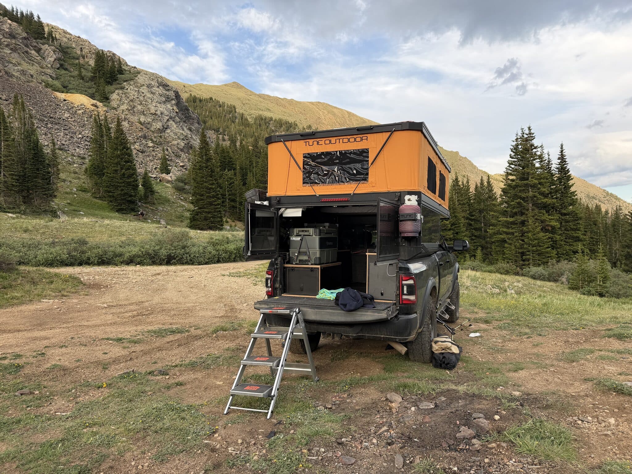 The truck camper parked in a high alpine valley with the orange pop-top raised, tailgate down with folding steps, and evergreen slopes behind.