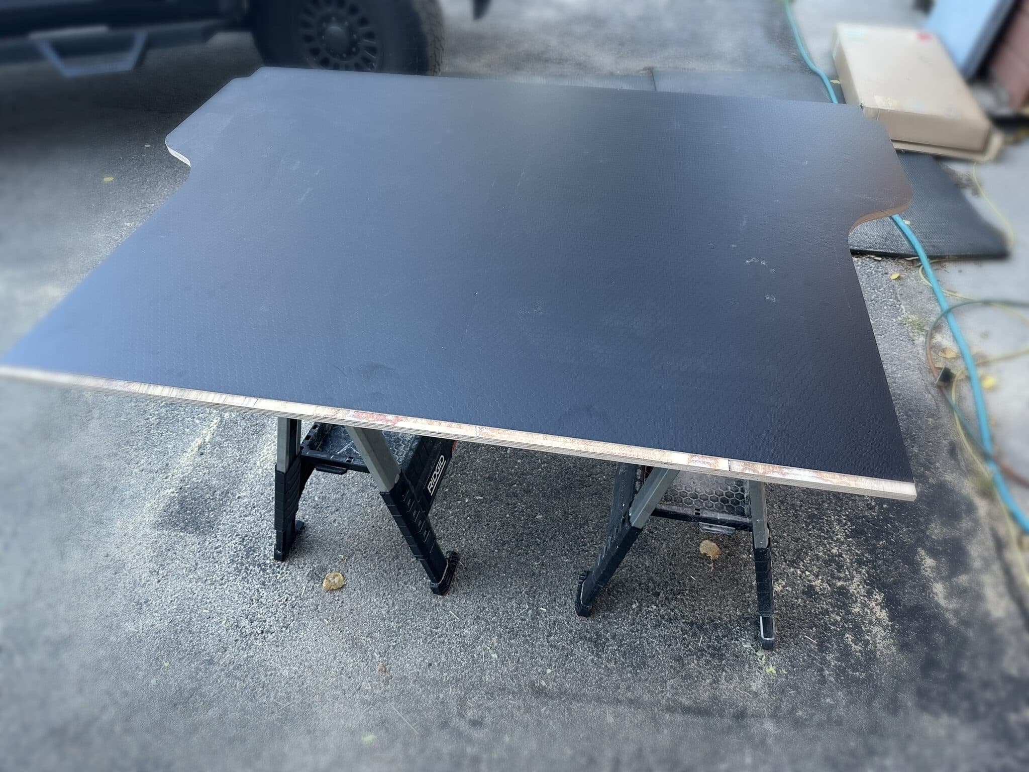 T-shaped plywood deck panel faced in black hex laminate resting on two folding sawhorses in the driveway, notched to fit around the truck bed's wheel wells.