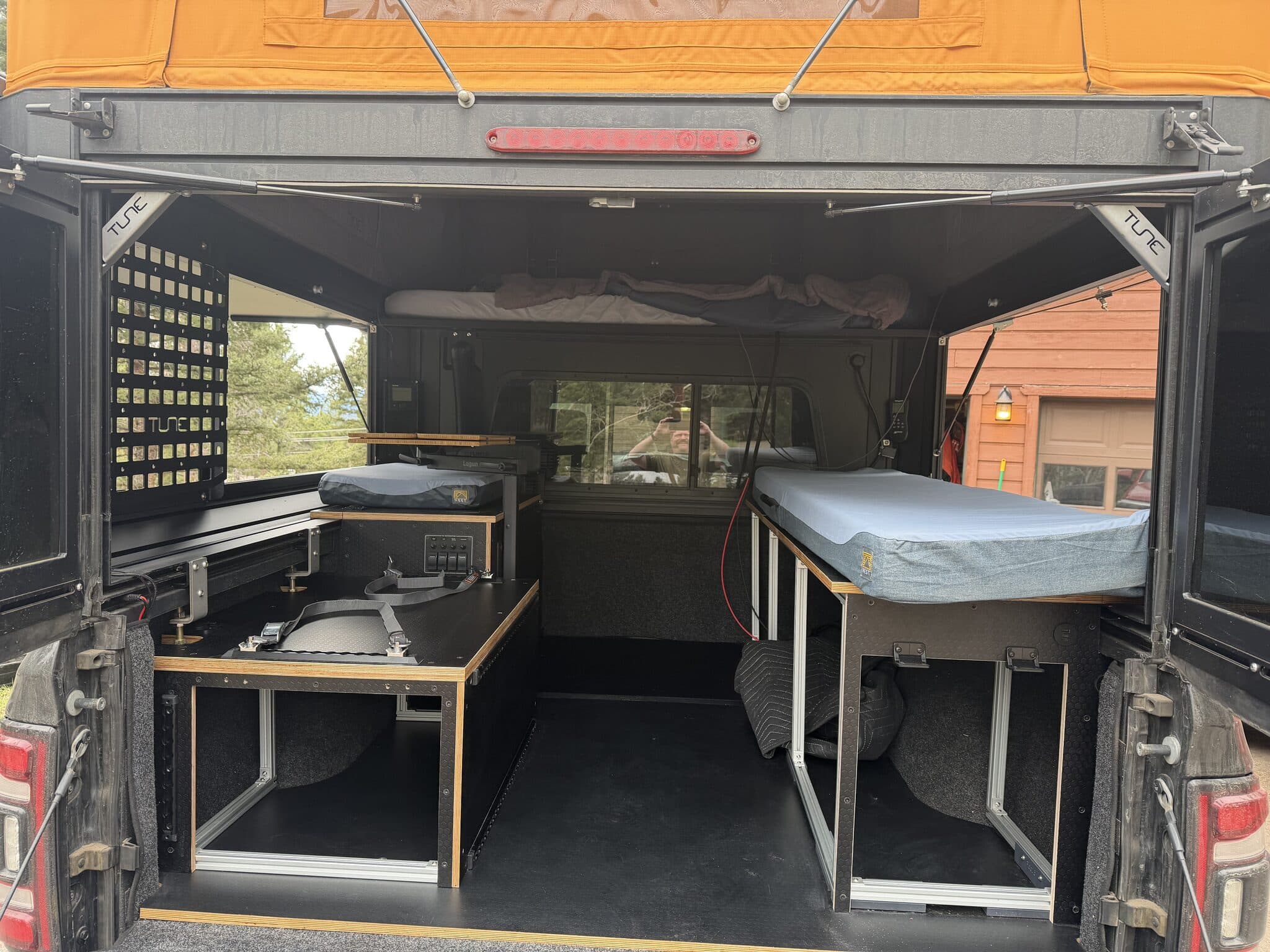 Rear of the camper with both doors open showing the finished build: bench modules on each side, mattress platforms, molle panel, and switch panel installed.