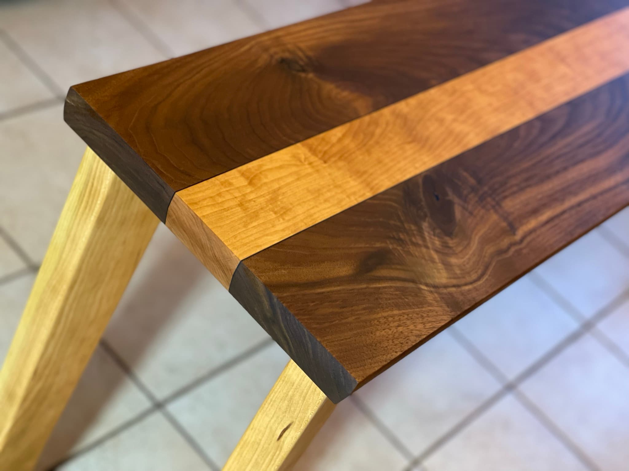 Three-quarter close-up of the table end: chamfered end-grain shows the walnut-ebony-maple-ebony-walnut sandwich, two tapered maple legs visible underneath.