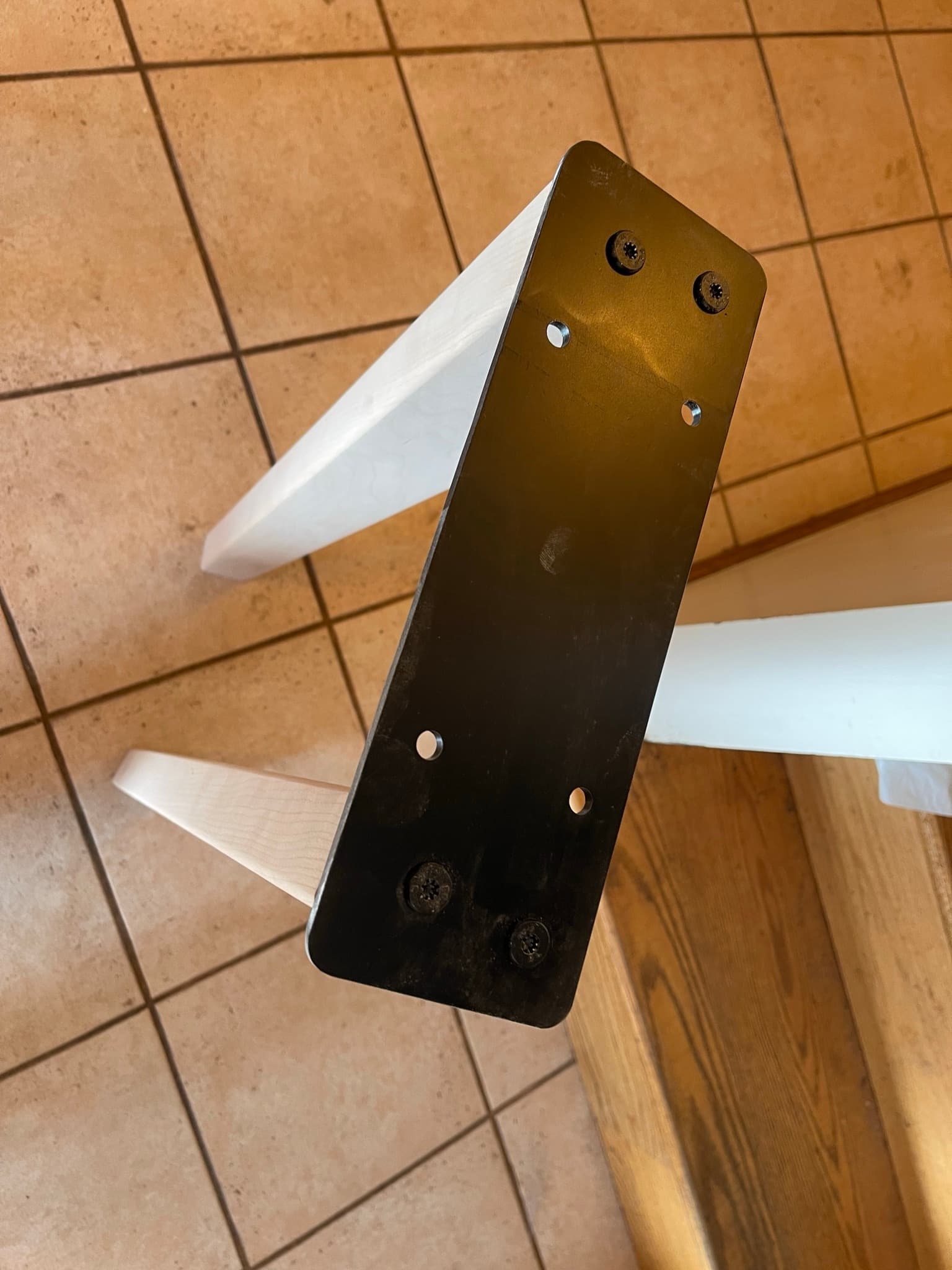 Two maple legs bolted to the blackened-steel structural plate, the assembly upside down on tile floor.